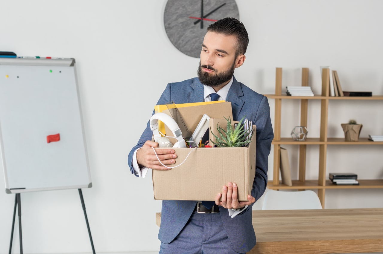 Man in business attire holding box of desk supplies standing in front of emply desk.