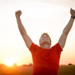 Man in red tshirt with arm raised in happiness with sun setting in background.