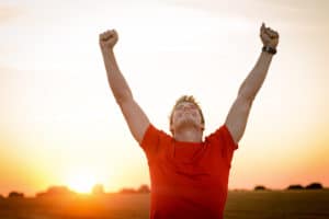 Man in red tshirt with arm raised in happiness with sun setting in background.
