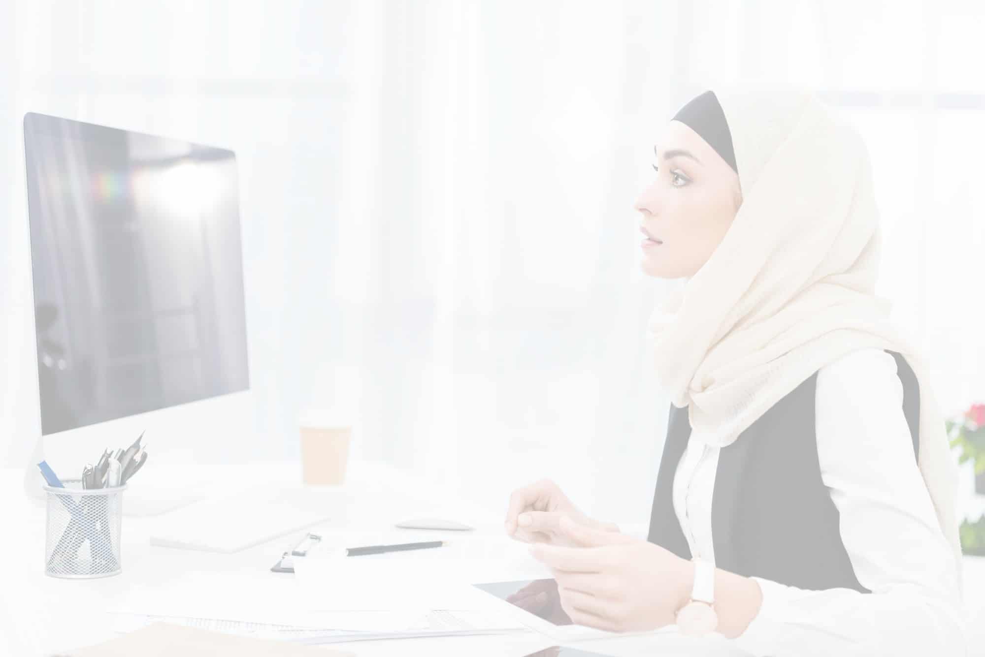 On the right, a professional Muslim woman sitting at her desk looking at computer monitor on the left.