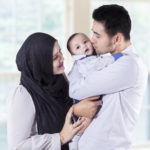 Muslim family kiss their baby while standing near the window at home.