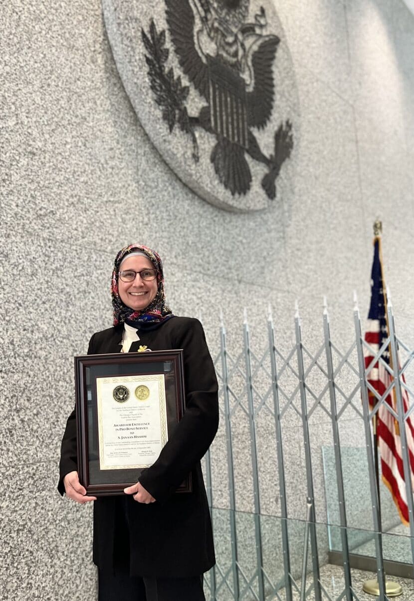 Muslim female attorney holding award, dressed in business formal wand red hijab, with American flag over her shoulder and the District Court Seal hanging on a wall behind her.