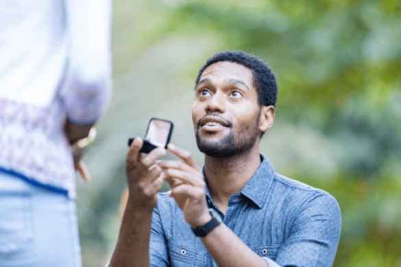 Man on knee with ring box open and looking up at woman with hopeful expression.