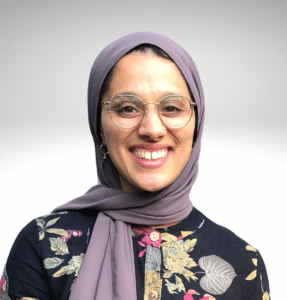 Sophia Akbar wearing mauve hijab, eyeglasses, black with floral print shirt against a gray to white background.